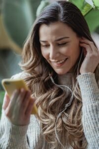 Young content female in casual sweater browsing modern mobile phone and listening to good music via earphones in living room